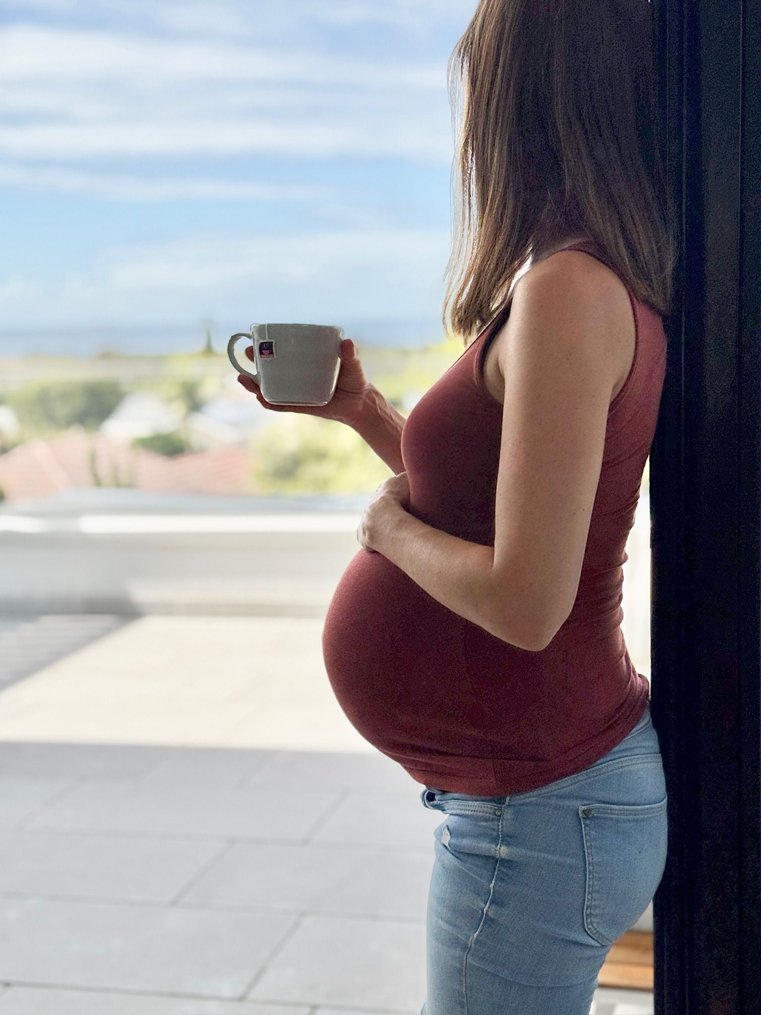 Pregnant woman holding a cup of Hottea Mama pregnancy tea while leaning against door frame, looking out at Sydney skyline