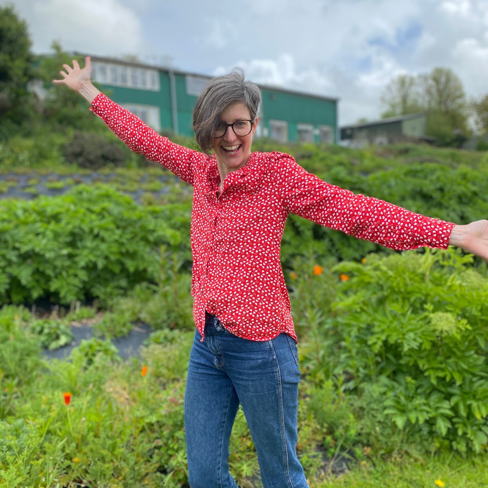 Women in red shirt stood in herb garden with arms open, looking at raspberry leaf tea and feungreek tea plants