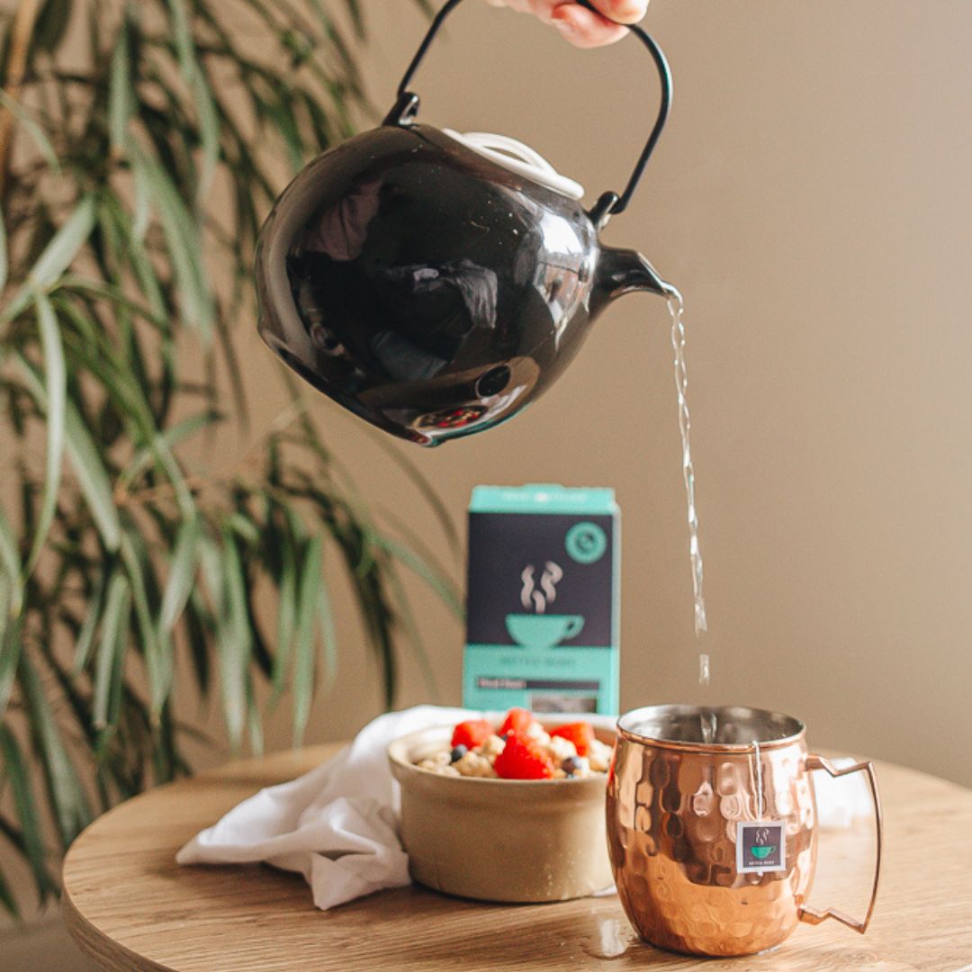 Black tea pot pouring hot water onto a HotTea Mama Head Start tea bag in a copper mug, with fresh fruit in a bowl behind.