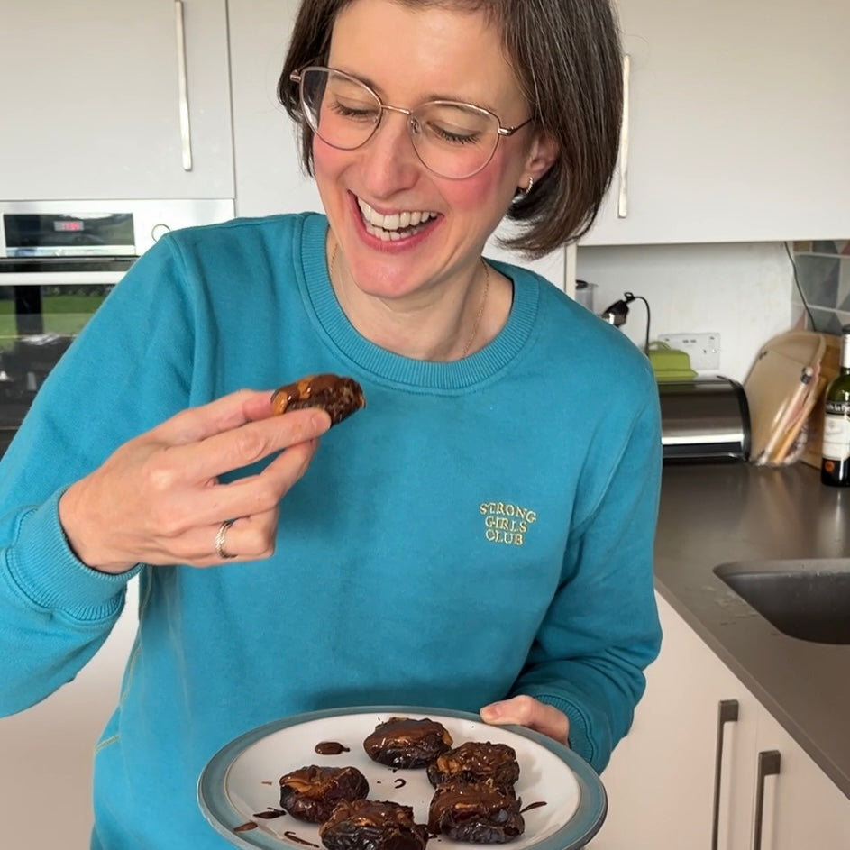 Brunette lady in turquoise jumper in kitchen eating dates that are stuffed with nut butter and covered in chocolate