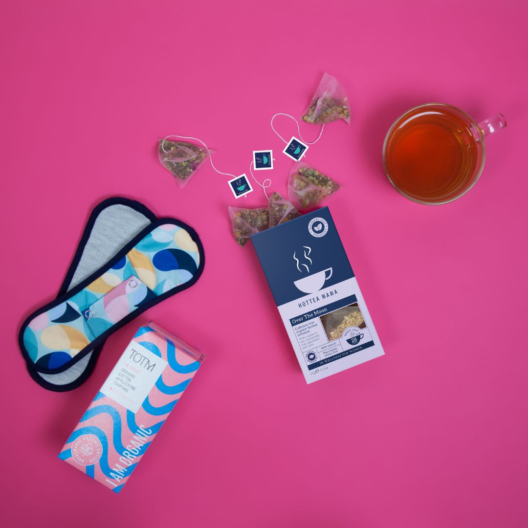 An open pack of Over The Moon period support herbal tea, on a pink background with a brewed cup of tea and whole leaf tea bags spilling out.  Reusable period pads and organic tampons lay next to the tea.