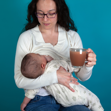 Mum breastfeeding her baby on a turquoise background, she is holding a glass cup with lactation hot chocolate in it