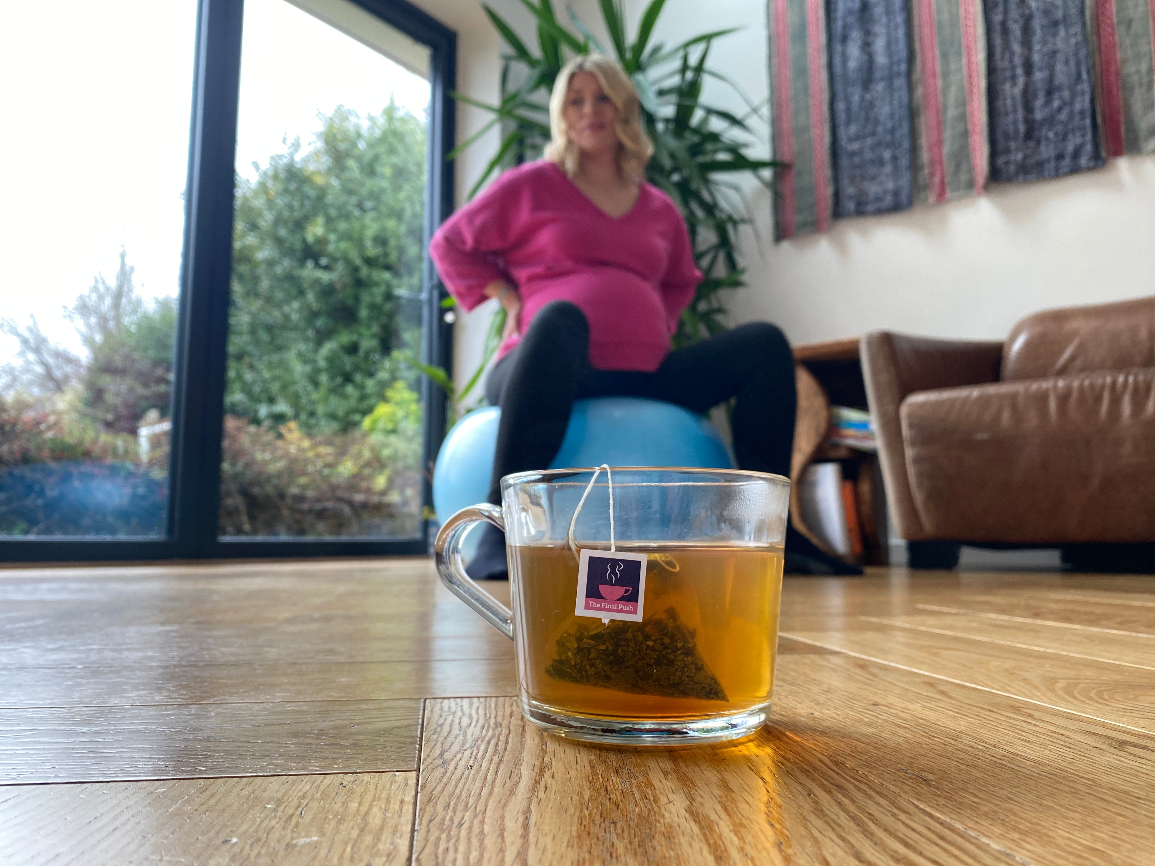 Pregnant lady in pink top, sitting on a yoga ball in black leggings, with a cup of HotTea Mama The Final Push raspberry leaf tea brewed in a glass cup in the foreground