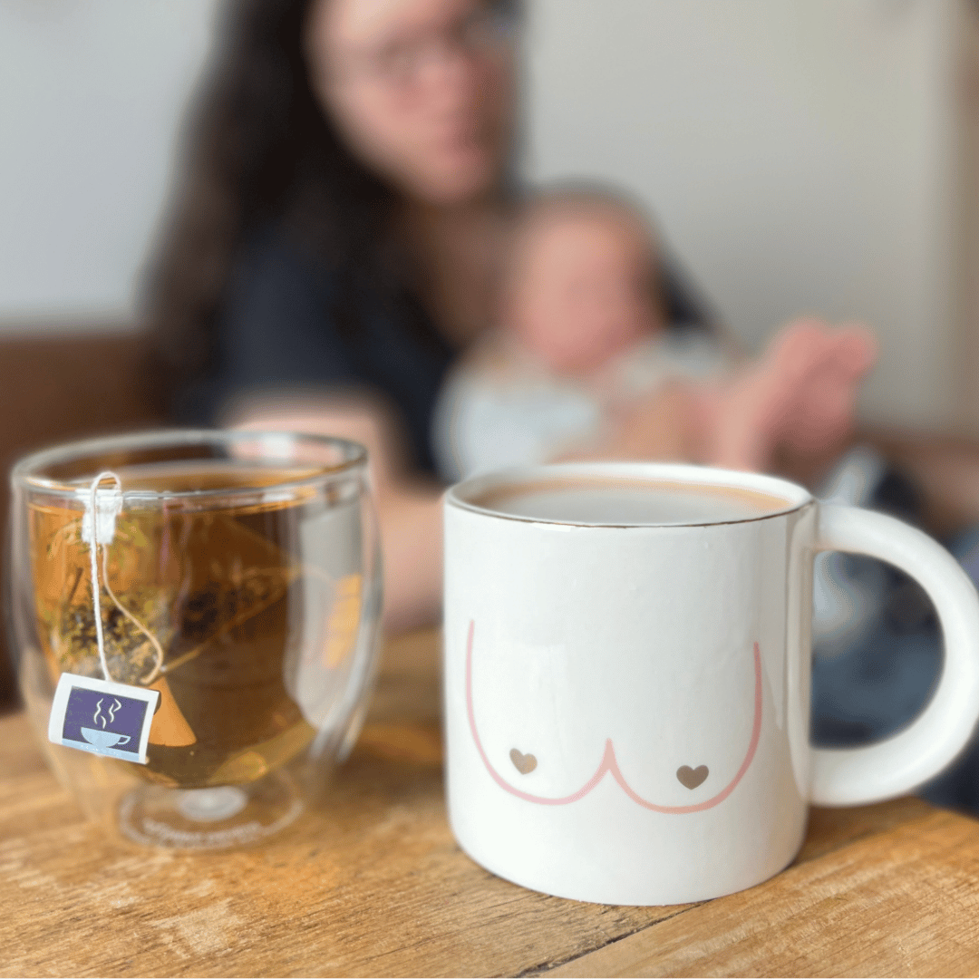 Nursing mother with baby in background, with HotTea Mama milk's up tea in glass cup in foreground and breastfeeding mug