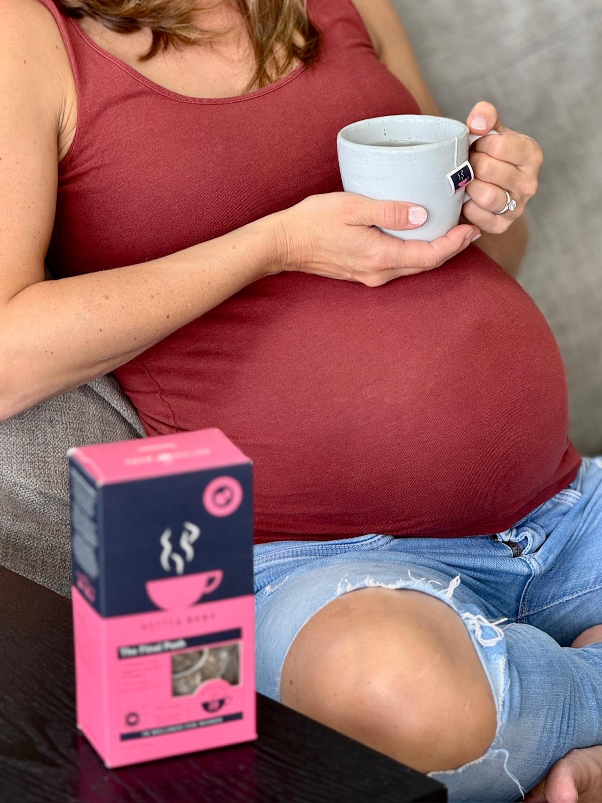 Pregnant lady in jeans and maroon vest top, holding a cup of HotTea Mama The Final Push raspberry leaf tea on her bump, with a pack of the tea on table next to the grey sofa she is sat on