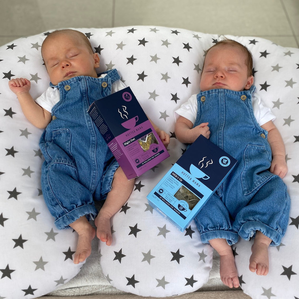 Twin babies in matching denim dungarees, lying on a feeding pillow next to a Night Owl Sleepy tea box and a Milk's Up breastfeeding support tea box