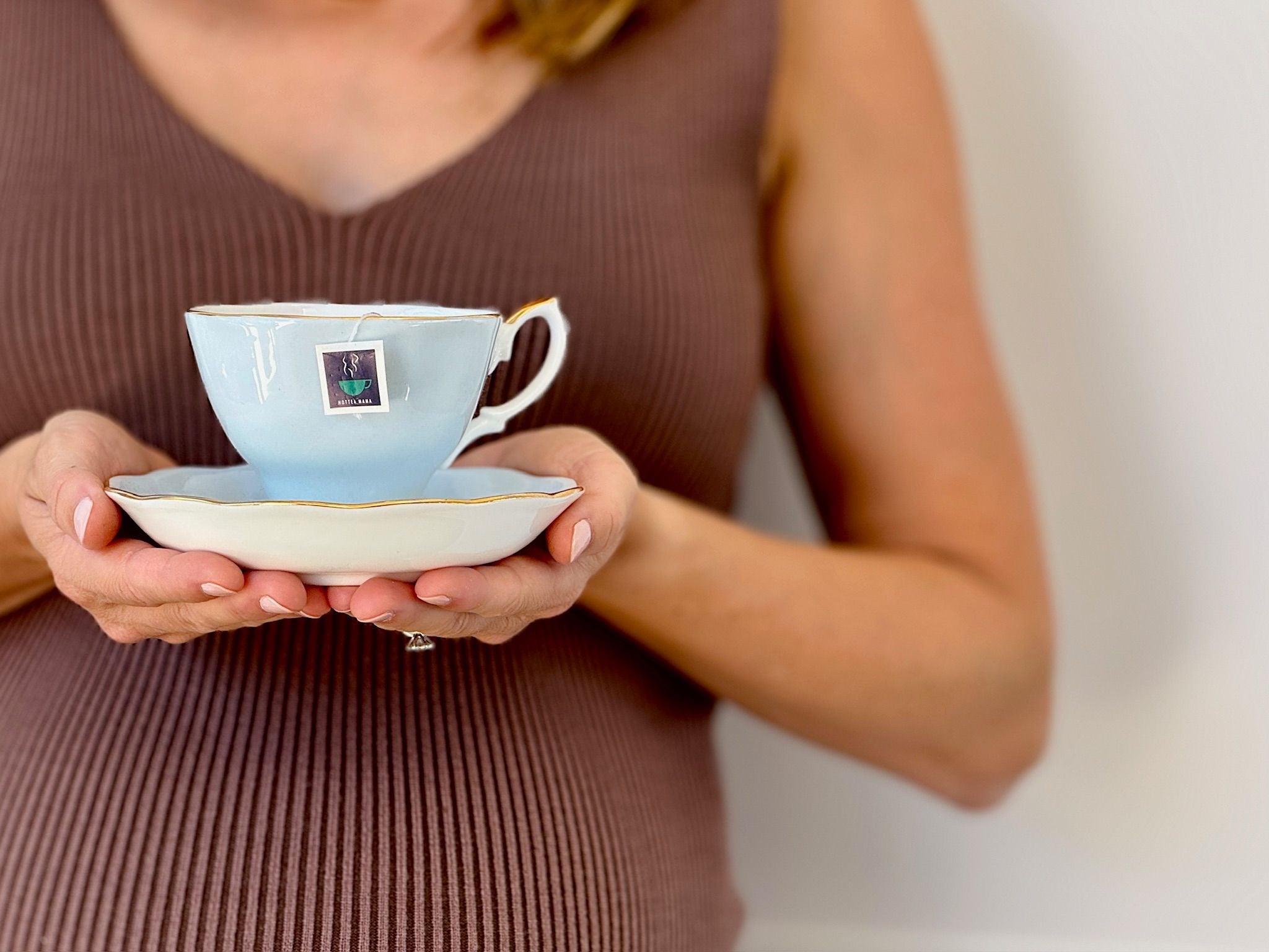 Woman in brown vest top holding a blue tea cup and saucer with HotTea Mama tea bag tag showing