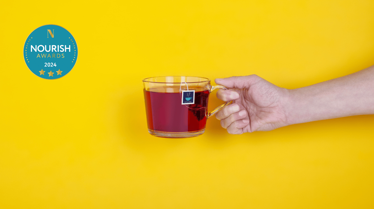 Hand holding a glass cup of get up and glow fruit tea, with a HotTea Mama tag, against a yellow background with the Nourish Awards 2024 3 gold star award logo