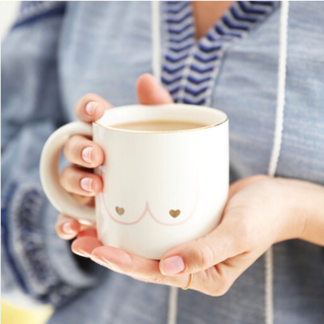 White porcelain mug with boobies on it, held in a ladies hand who is wearing a blue breastfeeding top