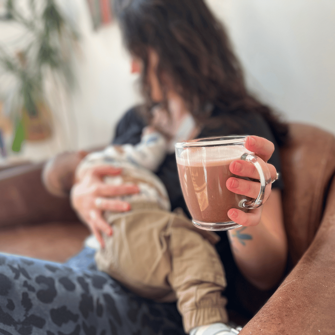 Mum holding a cup of HotTea Mama Go With The Flow Hot Chocolate whilst breastfeeding her baby on a brown sofa